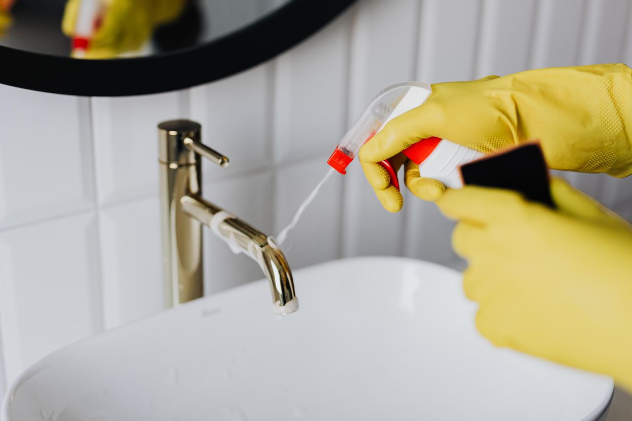 hero-img-01 From above anonymous housekeeper in yellow rubber gloves spraying cleaner to chrome water faucet in bathroom